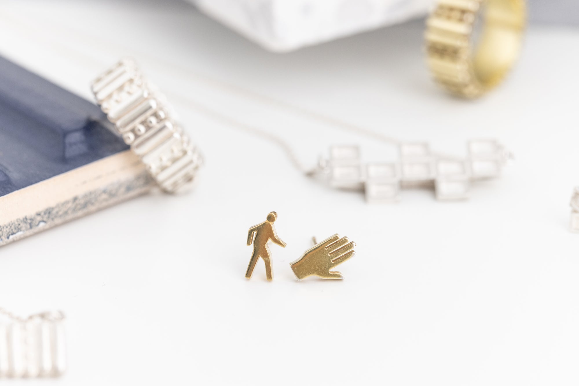 Close-up of 18k gold vermeil Crosswalk studs designed as walk sign human and hand icons, with Genji block ring on the left and concrete waffle necklace on the right in soft focus background.