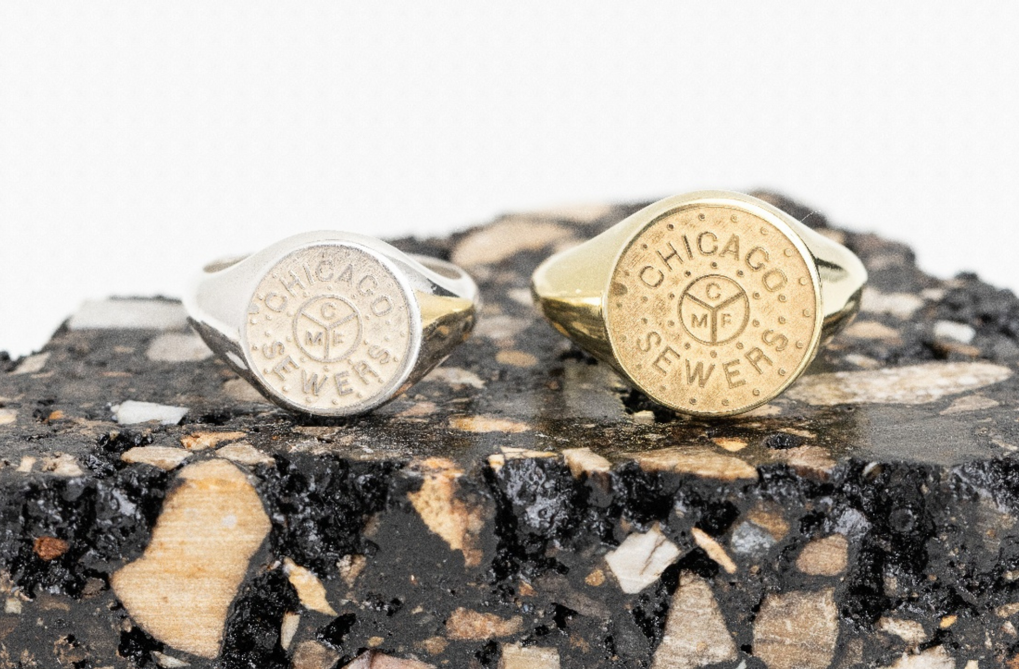 Sterling silver and 18k gold vermeil Chicago Mini Manhole rings displayed on a textured chunk of asphalt, highlighting urban-inspired jewelry design.
